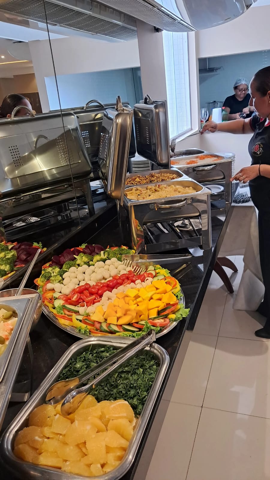 buffet de restaurante self-service com salada colorida em prato grande, gropas de inox e funcionária servindo