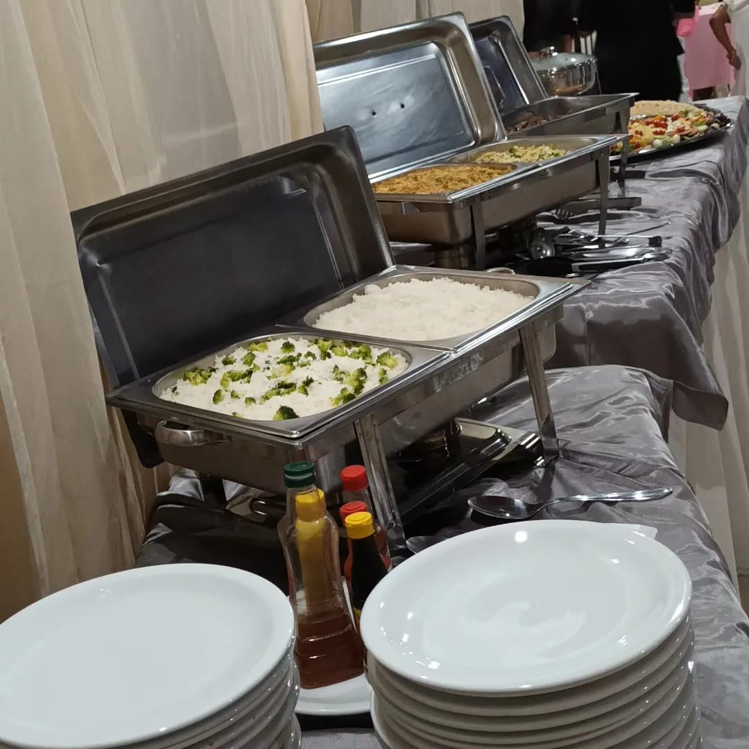 buffet de comida com rechaçês de inox, arroz com brócolis, pratos brancos empilhados e garrafas de molho sobre