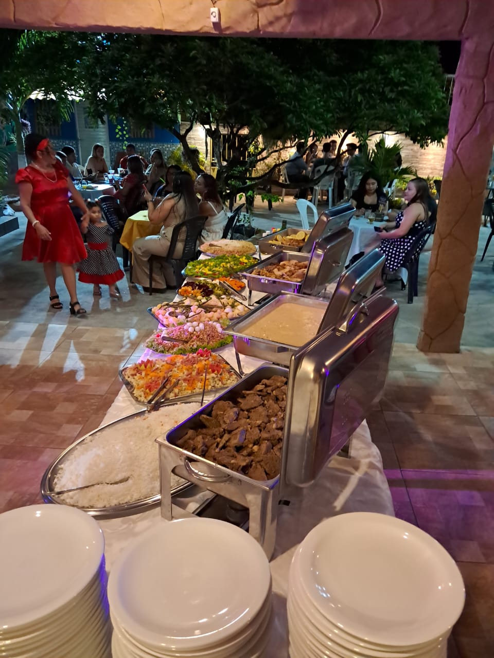 buffet de festa ao ar livre à noite, com rechaçês de metal, saladas, arroz, carnes e pilhas de pratos brancos,