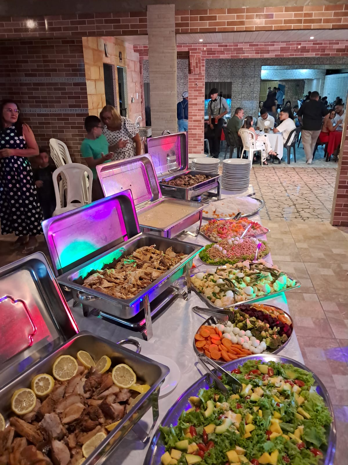 mesa de bufê de evento com rechauds de carnes, arroz e travessas de saladas variadas, salão cheio ao fundo