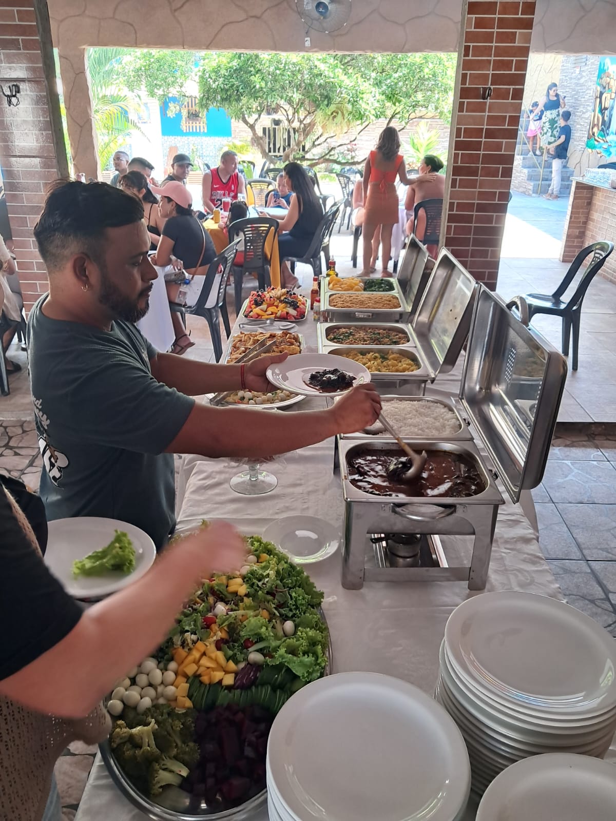 buffet de festa com garçom servindo comida em rechauds de inox, salada colorida no primeiro plano e convidados