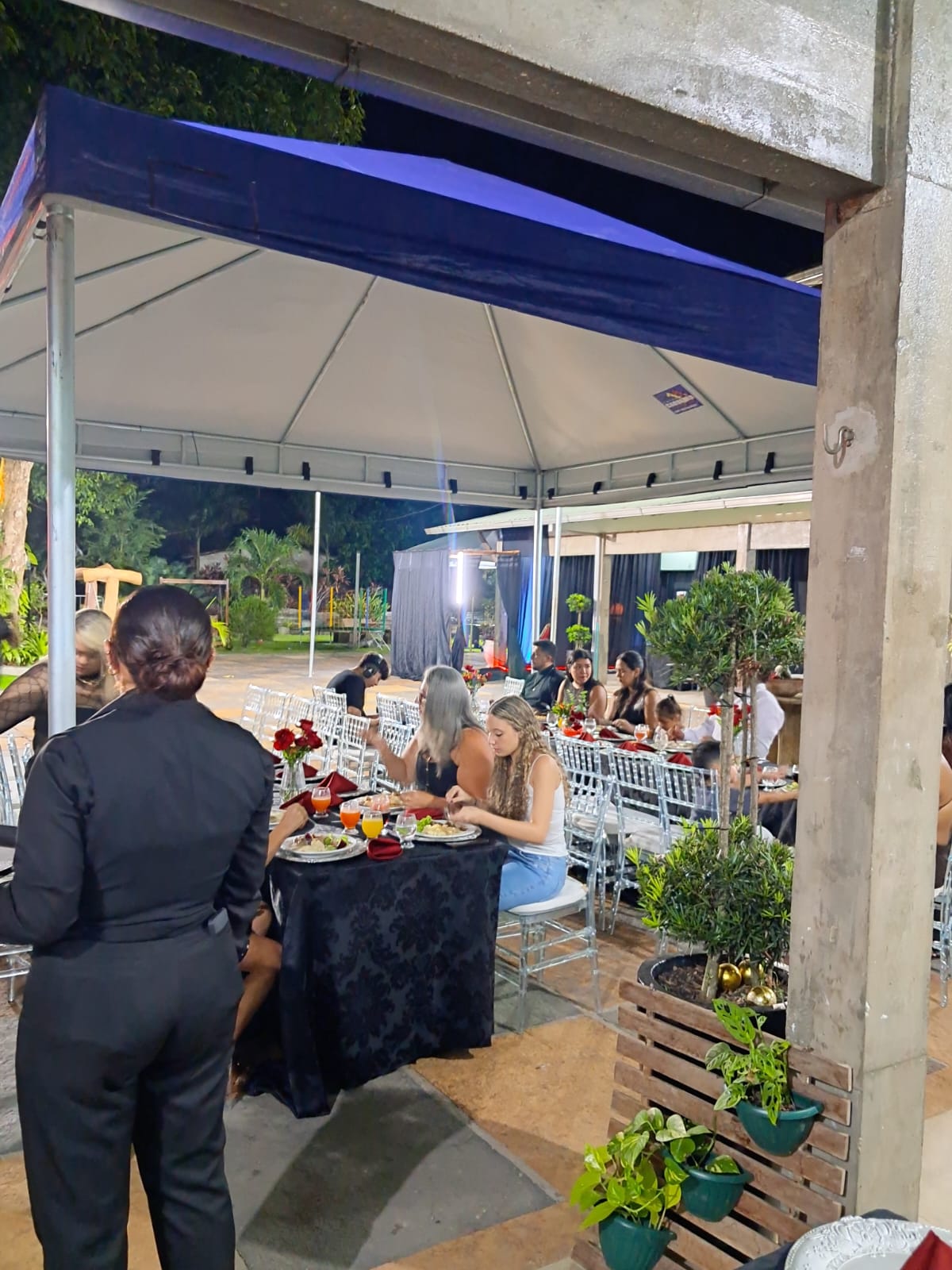 evento noturno em salão coberto com tenda azul, convidados sentados em mesas com toalhas pretas e garçonete de