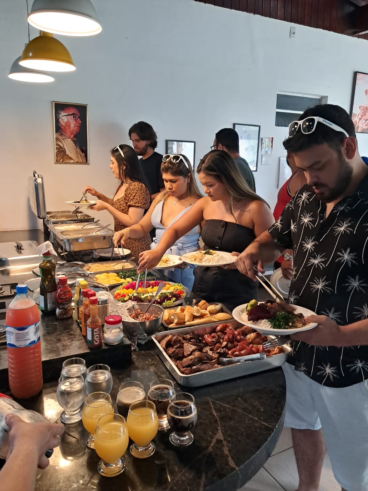 pessoas se servindo em um buffet self service com carnes, saladas, frutas e sucos em uma bancada de granito