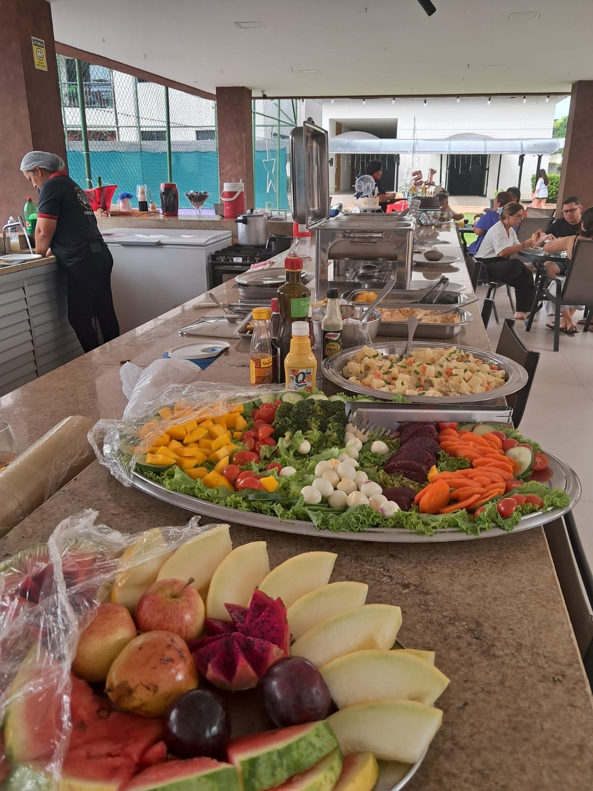 buffet de saladas e frutas em bandejas de inox sobre um balcão de granito, com funcionária ao fundo e pessoas 